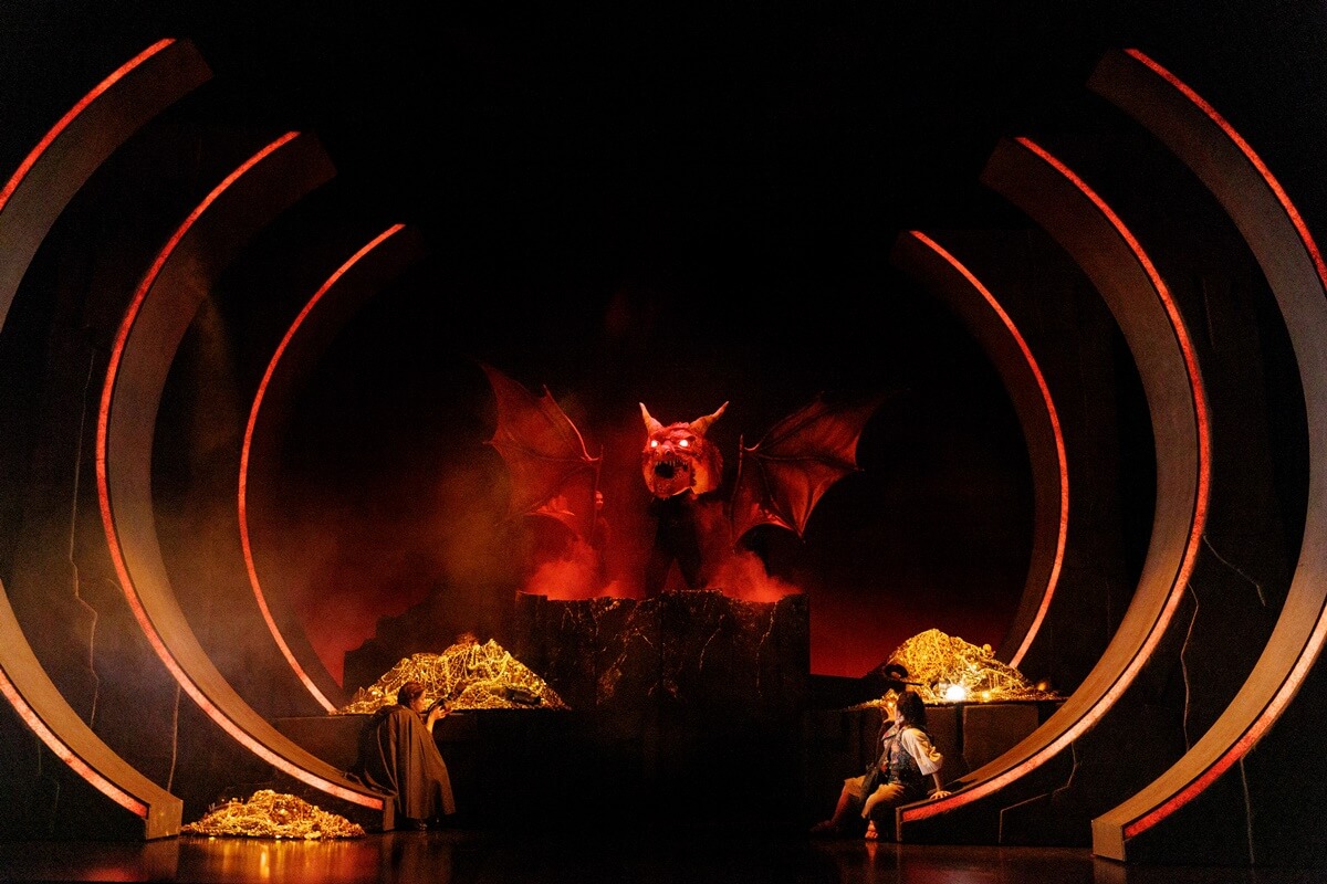 L-R: Sara-Jeanne Hosie as Old Took, Heidi Damayo, Tim Campbell as Smaug and Richard Lee as Bilbo Baggins, The Hobbit. Stratford Theatre Festival 2026 (Photo: David Hou) A large dragon figure stands atop gold treasure inside a circular stage set.