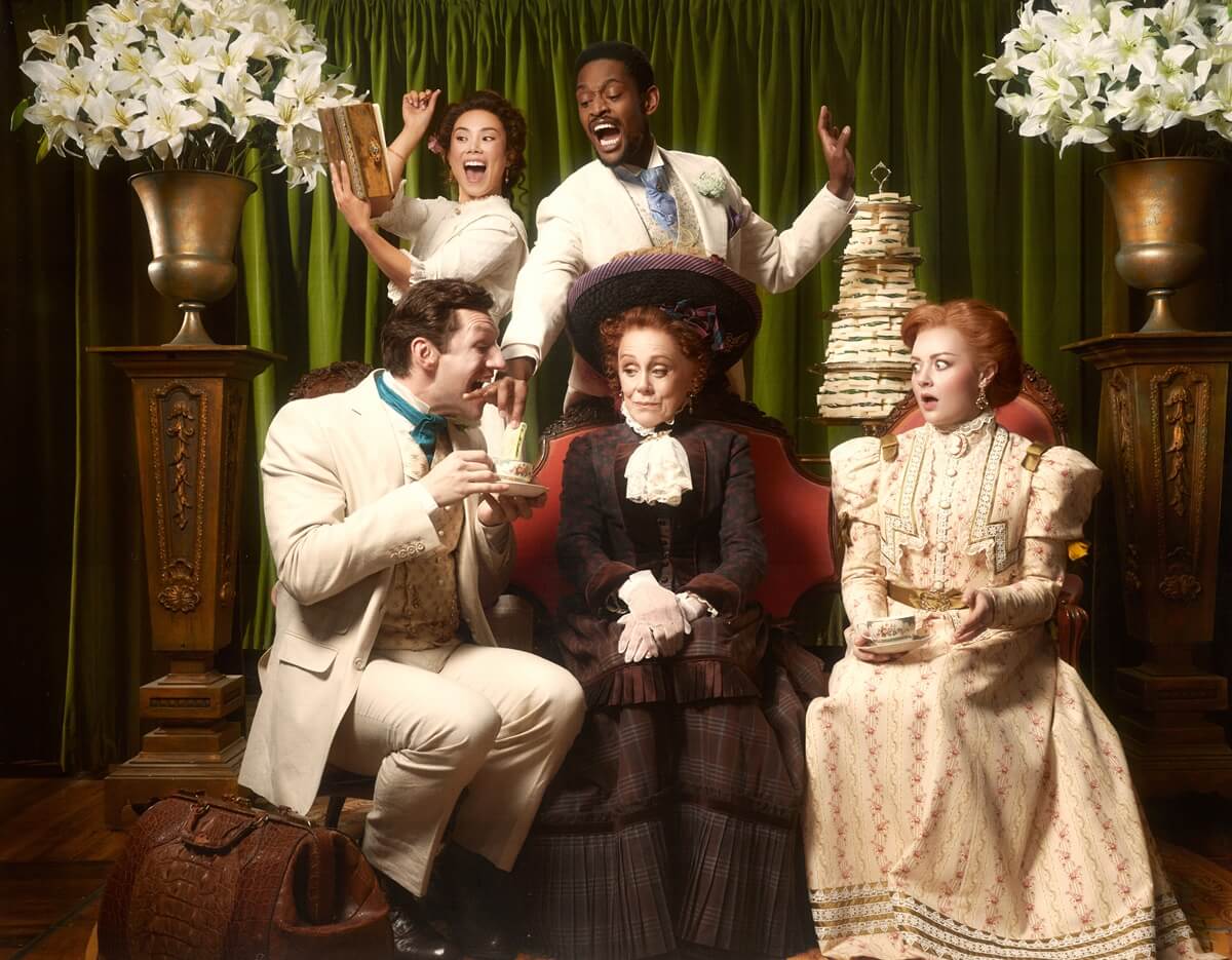 Six people in elegant vintage-style clothing pose in front of a green curtain, surrounded by large floral arrangements; some hold teacups, books, and a tiered stack of cucumber tea sandwiches. L-R: Joe Perry as John Worthington, Marissa Orjalo as Cecily Cardew, Christopher Allen as Algernon Moncrieff, Fiona Reid as Lady Bracknell and Allison Lynch as Gwendolen Fairfax, The Importance of Being Earnest. Stratford 2026 (Photo: Dariane Sanche)